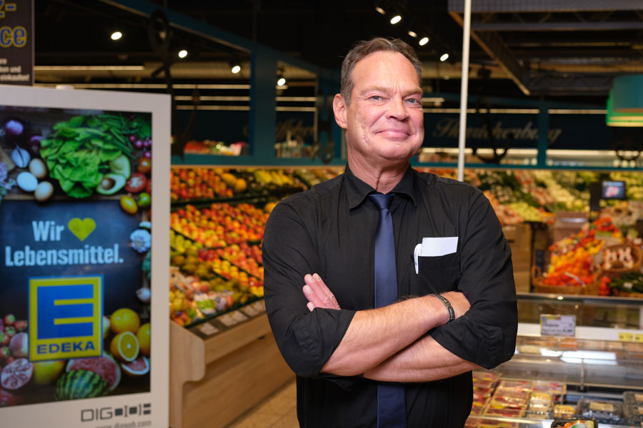 Inhaber im Supermarkt steht mit verschränkten Armen vor der Obst- und Gemüseabteilung neben Edeka-Werbeschild.