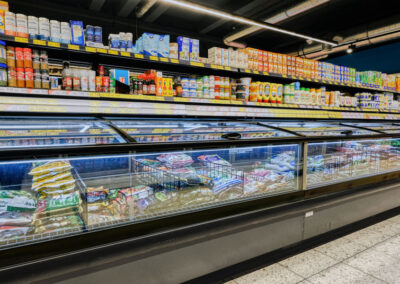 Tiefkühltruhe im Supermarkt mit gefrorenen Produkten, darüber Regale mit Gewürzen und Konserven.
